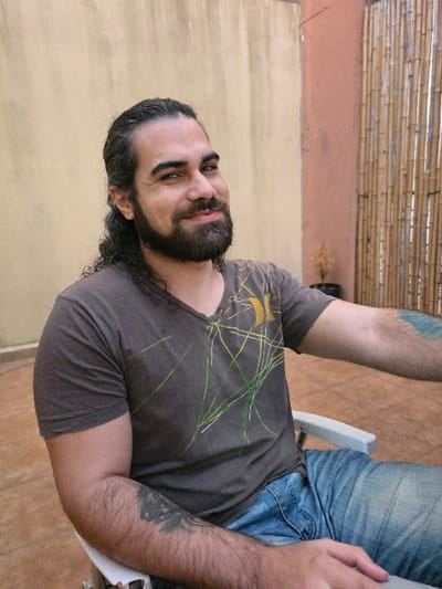 The image displays an adult male seated outdoors in Villa San Andrés, Argentina. The subject has dark, long, curly hair pulled back, a full dark beard and mustache, and is smiling towards the camera. He wears a dark grey V-neck t-shirt featuring yellow and green abstract linear designs, and light blue denim jeans. A dark tattoo is visible on his left forearm, and another on his right upper arm. He sits in a light-colored outdoor chair. The background consists of a plain light beige wall on the left, an orange-red wall on the right, and a vertical bamboo screen attached to the orange-red wall. A small, withered plant in a black pot is positioned on the ground between the walls. The visible ground area is covered with light brown tiled flooring.