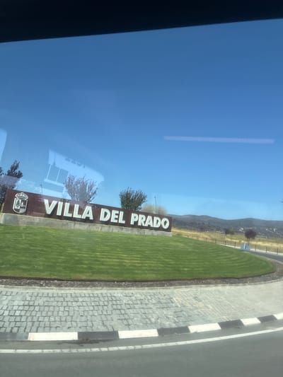 A sign that reads "VILLA DEL PRADO" is positioned in front of a grassy hill. The sign is brown with white lettering and a crest on the left side. The crest is white and depicts a crown on top of a shield with a castle and a tree inside. The grassy hill is well-maintained and slopes upwards behind the sign. In the background, there are trees and a dry, hilly landscape under a clear blue sky. A paved road curves in front of the grassy hill, with a sidewalk made of grey cobblestones. The scene appears to be an entrance to the town of Villa del Prado, Spain. The image is captured through a window, with reflections of buildings visible in the upper portion of the frame.