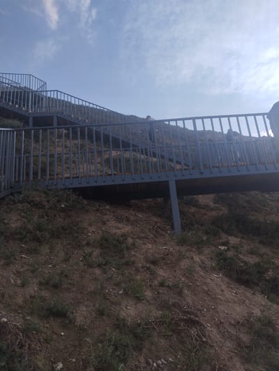 An outdoor, low-angle, wide shot captures a multi-tiered metal staircase ascending a barren, hilly slope under a bright, hazy sky in Naryn City, Kyrgyzstan. The staircase features a gray metal railing with vertical bars and wooden steps, supported by a dark metal structure. Several indistinct figures can be seen on the stairs, appearing as silhouettes against the sky, with one figure at the bottom of the visible portion of the stairs, and others further up. The ground is composed of dry, reddish-brown soil with sparse green vegetation and exposed roots. The sky is pale blue with wispy white clouds.