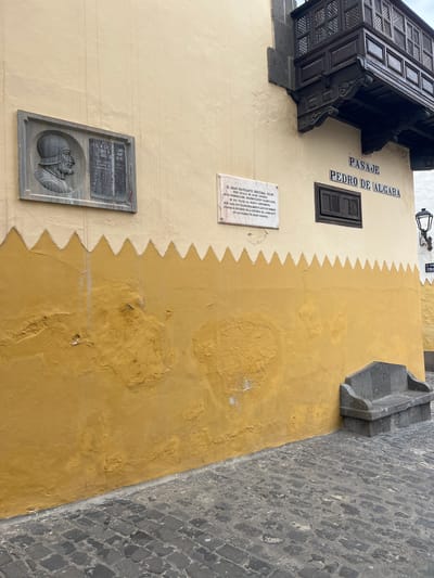 Here is a description of the image:
This is a ground-level shot of a building wall in Las Palmas De Gran Canaria, Spain. The wall has a decorative two-tone color scheme, with the top part being a lighter yellowish cream, and the lower portion being a darker mustard yellow. The demarcation line between these colors is zig-zagged.
The image features several plaques and architectural details attached to the wall:
1. A small, dark wooden window with a decorative exterior frame.
2. A black wooden balcony with intricate patterns of holes.
3. A white plaque with black lettering, containing historical information about Christopher Columbus' voyages to Gran Canaria.
4. A grey plaque featuring a relief of a person, possibly a historical figure or symbolic image.
5. A stone fixture extends from the wall, possibly a small balcony or architectural detail.
The words "PASAJE PEDRO DE ALGABA" are painted on the wall in black lettering, referring to the name of a local alleyway.
To the right of the wall, is a portion of the street, paved with grey cobblestones. The wall is clearly aged with some weathering and surface imperfections.