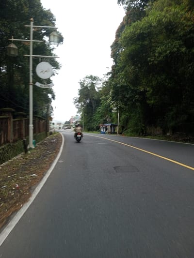 The scene depicts a curved asphalt road in Phoxay, Laos, under an overcast sky. The road extends from the foreground into the distance, gently curving to the left. A solid yellow line marks the center of the road, while a solid white line delineates the right edge of the paved surface.

On the left side, a low, ornate concrete wall runs parallel to the road, behind which dense green foliage is visible. A prominent, light-grey lamppost stands on the left, equipped with two decorative lanterns and a circular white sign bearing Arabic script ("العزيز") above a smaller sign reading "AL AZIZ". The area between the white line and the wall consists of dirt and sparse vegetation.

On the right side, a steep embankment covered in dense, tall green trees and thick vegetation borders the road. Further down, a small, light-blue and white roadside stall or shack is partially visible, nestled within the foliage.

One individual, wearing a helmet, is operating a dark-colored scooter or motorcycle, traveling away from the camera in the center-right lane of the road. No other persons are present. The road surface is dry, and the environment is characterized by lush greenery. Distant structures are faintly visible at the horizon.