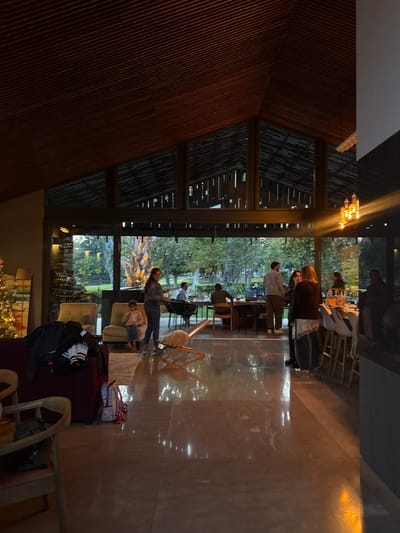 A large, dimly lit hall is filled with people. Tables are set for dining near the windows, while others stand around socializing. Outside, trees are visible through the expansive glass. The hall features wooden walls and a high, angular ceiling with hanging chandeliers. In the foreground, furniture is scattered around, suggesting a more relaxed lounging area.