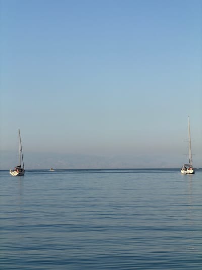 Two sailboats are sailing on a calm body of water under a clear sky. The location appears to be near Chianchitta, Italy, based on the coastal geography visible in the background. The sailboats are spaced apart with one in the foreground and the other in the distance. The water has subtle rippling patterns.