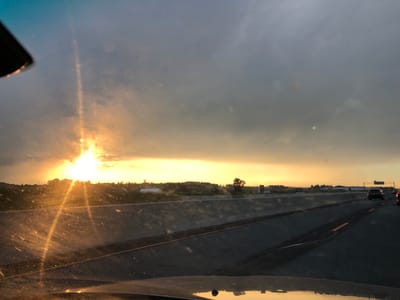 The image depicts a view from inside a vehicle, looking forward onto a highway at sunset. The sun is low on the horizon, casting a bright, warm glow across the sky and illuminating the clouds with shades of yellow and orange. Sunbeams stream through the windshield, which is speckled with dust or water droplets, creating a lens flare effect.
The highway stretches ahead, with a concrete barrier separating the lanes. The road surface is dark asphalt, marked with yellow lines. Several vehicles are visible on the highway, moving in the same direction as the viewer. A dark-colored SUV is prominent in the right lane, with another vehicle partially visible behind it.
In the background, beyond the highway and barrier, is a rural landscape. Fields and trees are silhouetted against the bright sky. Some man-made structures, possibly farm buildings or industrial facilities, are scattered across the landscape. The sky transitions from the warm sunset colors near the horizon to a cooler, grayish-blue towards the top of the frame, indicating a mix of clear sky and clouds.
The time of day is clearly sunset, given the position and color of the sun. The weather appears to be fair, with the clouds adding to the dramatic lighting. The presence of a highway with other vehicles suggests an ongoing journey or commute. The overall mood is serene and visually striking, emphasizing the beauty of the natural light. No visible text is present in the image. The location context is Bradford West Gwillimbury, Canada.