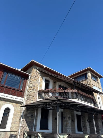 A multi-story stone building with a brown roof and dark wood accents stands against a clear blue sky. A thin black wire stretches diagonally across the sky. The building features multiple windows, some with dark shutters, and balconies adorned with flower boxes. A covered patio with black metal supports extends from the lower level. The overall scene suggests a clear, sunny day. The image is captured from a low angle, looking up towards the building.