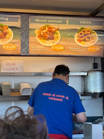 The image depicts an outdoor food stall in Zandvoort, Netherlands. The stall features a menu board displaying three seafood options: "eling", "Mixed seafood", and "Inktvis". Each option is accompanied by a price in Euros and a brief description. Below the menu board, a sign indicates the stall is closed at 18:00. A man in a blue t-shirt with orange lettering saying "REGEL IK VOOR JE, VRIEND!!!" is visible, facing away from the camera. He appears to be working at the stall. Various food containers and serving dishes are stacked on shelves behind him. The overall environment suggests a casual dining establishment, likely near a beach given the backdrop of the sea on the menu.
