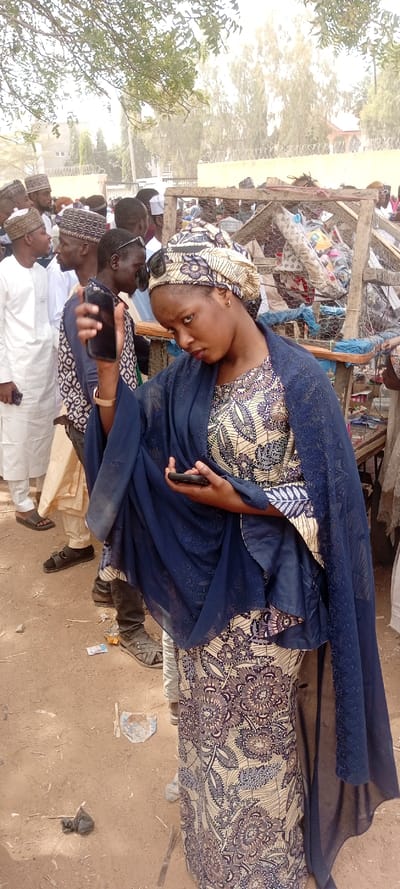 The image depicts a public outdoor gathering, likely a market, in the city of Sani Mai Nagge, Nigeria. In the foreground, a woman faces slightly right of the camera, looking towards the viewer with a neutral expression and her head tilted. She wears a cream-colored headwrap with blue and brown patterns, matching a long dress. A dark blue, semi-sheer veil or shawl covers her shoulders and drapes over her left arm. She holds a black smartphone in her left hand, which is slightly raised, and another black smartphone in her right hand, positioned near her hip.
Behind the woman, a crowd of individuals, primarily men, is visible. Many wear traditional Nigerian attire, including long white robes (tagiya) and various patterned caps. Some individuals wear sunglasses. To the right of the central woman, a makeshift stall constructed from wooden frames and chicken wire is visible, containing various items such as fabric and packaged goods, suggesting a retail or market environment.
The ground is unpaved, consisting of light brown dirt with scattered debris, including small pieces of paper and dark refuse. In the background, behind the crowd and stall, green foliage from trees is visible, partially obscuring distant light-colored buildings with security fencing topped with barbed wire. The lighting suggests daytime conditions.