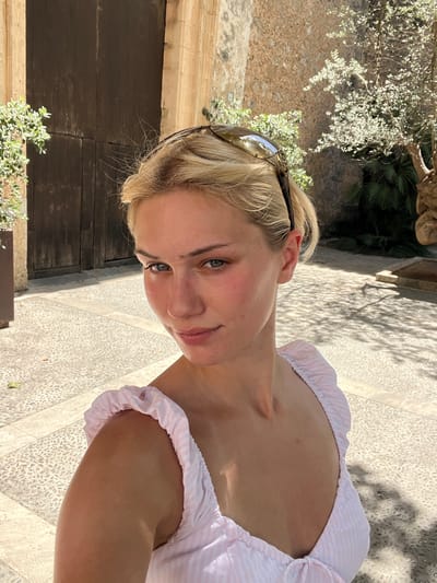 In Pollença, Spain, a young woman with blonde hair pulled back and sunglasses perched on her head poses for a selfie outdoors. She is wearing a light pink and white striped off-the-shoulder top. The background features a rustic stone wall and a large, dark wooden door, with some greenery and shadows from trees visible. The ground is paved with small, light-colored stones. The lighting suggests it is daytime, likely sunny, with strong shadows.