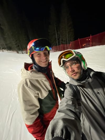 At night, on a snow-covered ski slope in Jahorina, Bosnia and Herzegovina, two adult males are posing for a selfie. The individual on the left wears a red ski helmet with blue and yellow mirrored goggles, a black neck gaiter, and a multi-colored winter jacket (beige, dark green, and red sections). He is smiling at the camera, with a snowboard partially visible on his left side. The individual on the right wears a white and green patterned ski helmet with orange and yellow goggles, clear-rimmed glasses underneath, a black neck gaiter, and a grey winter jacket. He holds the camera, capturing the image, and looks at the camera with a neutral expression. The background features dark evergreen trees, a red safety net along the right side of the slope, and faint artificial lights in the distance. The scene is illuminated by artificial lighting, indicating night skiing conditions.