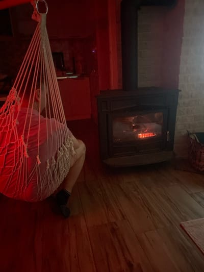 An indoor scene in Palež, Montenegro, displays a seated individual in a white macrame-style hanging chair, viewed from behind. The person wears a light-colored upper garment and dark socks, facing a dark-toned, corner-positioned wood-burning stove with visible flames behind its glass door. The room's floor is composed of wooden planks, and a segment of a white brick wall is visible behind the stove. The ambient lighting throughout the space exhibits a prominent reddish hue. In the distant background, a dark object resembling a screen is faintly discernible. A dark woven basket and a light-colored rectangular rug are partially visible on the floor to the right of the stove.