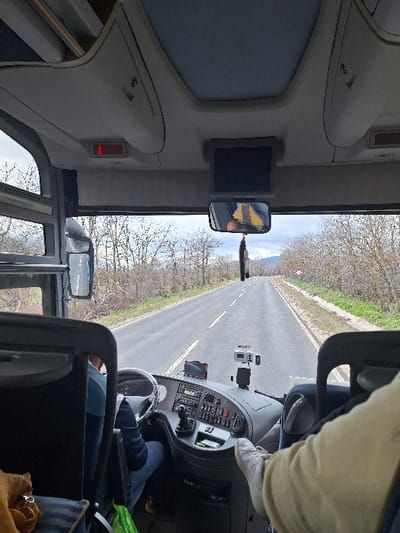The image depicts the interior perspective from a moving bus or coach in Gabarevo, Bulgaria, looking forward through the windshield onto a two-lane asphalt road. The road is lined on both sides by leafless deciduous trees and some green undergrowth, indicative of late autumn, winter, or early spring. An overcast sky is visible above.
In the foreground, the lower legs and feet of a passenger, wearing light-colored pants and beige shoes, are elevated and resting on the right side of the vehicle's interior, near the dashboard. To the left, the bus driver is partially visible, wearing a blue long-sleeved shirt and blue jeans, with hands on the steering wheel, actively operating the vehicle. The dashboard features a steering wheel, a gear selector, instrument clusters, various buttons, a mounted GPS device, and an action camera.
Overhead, a small black monitor is centrally mounted, below which a rearview mirror reflects a yellow object and a partial human head. Storage compartments and a red illuminated indicator light are visible on the overhead panel. A green shopping bag and a brown textile item are situated on the floor or seat to the lower left. In the distance on the right side of the road, a circular red-and-white prohibitory road sign is present. Distant mountains are faintly visible under the overcast sky.