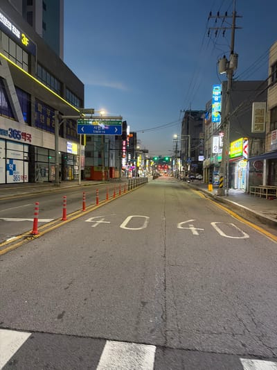 The image shows a street scene in Uijeongbu-si, South Korea, at dusk. The street is paved and has yellow lane markings. Traffic cones are placed along the right side of the road. White numbers "40" are painted on the road surface, indicating a speed limit. Buildings line both sides of the street, featuring various signs and illuminated windows. A traffic light is visible in the distance, showing a green light. The sky is a deep blue, suggesting twilight. There are no people clearly visible in the foreground.