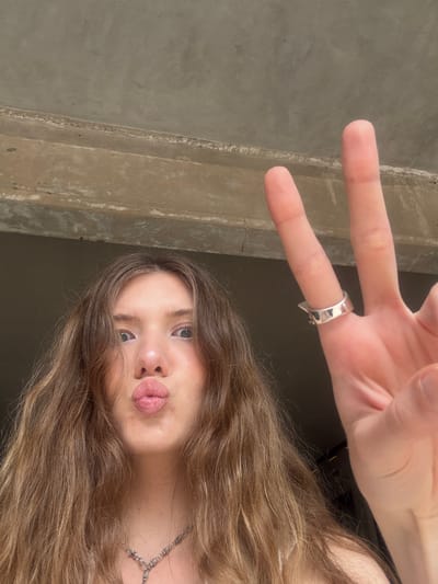 A young Caucasian woman with light brown, wavy hair is holding up her hand in a peace sign. She has light skin, blue eyes, and is pouting her lips. She is wearing a silver ring on one of her fingers and a silver necklace. The background appears to be a concrete structure, possibly a building. The picture was taken in Hongkou District, China.