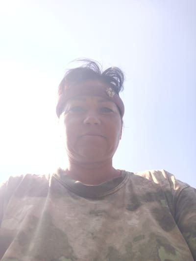 A woman is pictured from a low angle, looking upwards. She has dark hair pulled back and is wearing a reddish-brown headband adorned with small pearl-like beads. Her shirt is a camouflage pattern in shades of green and tan. The background is a bright, clear sky, suggesting an outdoor setting. The lighting indicates it is daytime, possibly sunny, with the sun positioned high and to the left of the frame, causing some overexposure in that area. The image was taken in Zalesovo, Russia.