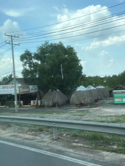 This is a landscape, street-level shot of a roadside establishment, likely a restaurant, in Long Thành, Vietnam. The image is taken from a moving vehicle, indicated by the motion blur and the perspective. The establishment features several thatched-roof huts, resembling small gazebos or dining areas. A prominent green building with signage is also visible.
Several signs with Vietnamese text are present. One sign reads "BIA SAIGON BAO LINH," indicating a beer brand and likely the name of the establishment. Another sign mentions "QUÁN ĂN BAO LINH," translating to "Bao Linh Eatery." Further text on a lower sign mentions "ĐẶC SẢN: GÀ TA" which means "Specialties: Free-range chicken," and "TIỆC CƯỚI HƠI LIÊN HOAN," suggesting it caters to parties and banquets.
The scene appears to be set during the daytime, with a bright sky and scattered clouds. The weather seems fair. There are no people clearly visible in the image. The foreground shows a grey metal guardrail and a paved road with a white line. The background consists of lush green trees and vegetation, characteristic of a rural or semi-rural setting in Vietnam. The overall impression is of a casual, possibly traditional, dining spot along a busy route.