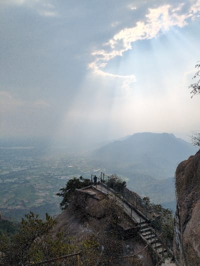 The image presents a panoramic view from a high, rocky elevation overlooking Kadaladi, India. In the midground, a dark grey stone staircase, equipped with dark metal railings, is constructed into a steep, rocky incline. The stairs lead to a small, flat viewing platform where a single individual stands silhouetted against the bright background, observing the expansive vista. Sparse dark green vegetation clings to the rocky terrain surrounding the stairs and platform. A large, dark rock face partially frames the right side of the scene. The background features a vast valley displaying a patchwork of green agricultural fields and scattered structures, fading into hazy blue-grey distant mountains. A body of water is faintly discernible in the far left background. The sky overhead is light grey, heavily clouded, with distinct crepuscular rays breaking through a cloud gap on the upper right, illuminating the atmospheric haze.