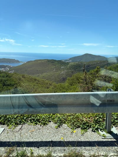 The image captures a scenic view of the coastline and verdant hills, presumably from an elevated position overlooking the city of Stanišići, Montenegro. In the foreground, a metal guardrail, typical of a roadside, spans across the bottom of the frame, with concrete and asphalt visible beneath it. A patch of green ivy grows along the edge of the road.

The middle ground is dominated by rolling, densely forested hills that descend towards the sea. Scattered structures, likely buildings within Stanišići, can be observed nestled amongst the trees in the valley. In the distance, the calm, blue expanse of the Adriatic Sea stretches towards the horizon, meeting a clear, bright blue sky. Wispy white clouds are present in the upper portion of the sky, and a faint contrail from an aircraft is visible. The overall impression is one of a bright, sunny day in a coastal mountainous region.