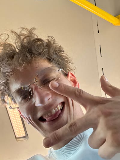 A low-angle, close-up selfie shows a person's face from the neck up. The person has curly brown hair and is wearing rimless glasses. Their right eye is squinted, and their tongue is sticking out. They are making a peace sign with their left hand. Behind them, a yellow pole is visible, along with a light fixture and a plain wall. The scene appears to be indoors, possibly on public transportation given the presence of the pole and light fixture. The location is not identifiable as Villa del Prado, Spain.
