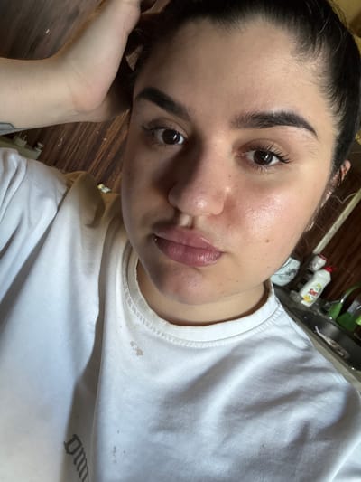 A young woman with dark hair pulled back is taking a selfie, resting her right arm on her head. She is wearing a white t-shirt. The background appears to be an indoor setting with wooden paneling on the wall and a kitchen counter with cleaning supplies visible. There are water stains and possibly food stains on her t-shirt. The image is taken from the chest up, focusing on her face and upper body. No specific location in Kadiivka, Ukraine is identifiable from the visual information.