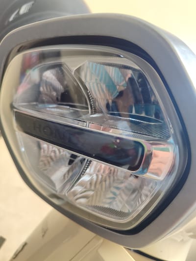 A close-up shot focuses on the headlight of a scooter, with the "HONDA" logo clearly visible on a chrome accent strip. The headlight features a multifaceted internal design, with clear plastic casing reflecting the surrounding environment, including what appears to be a ceiling light and patterned surfaces. The scooter itself is a light cream color, and the headlight is encased in a matte grey housing. The background is blurred, suggesting shallow depth of field. The image is cropped tightly on the headlight, emphasizing its details and the branding. The scene is indoors, with artificial lighting. There are no people or animals visible in the image.