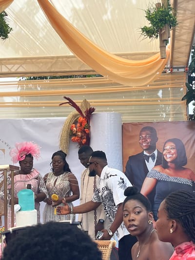 An outdoor event is depicted in Kumasi, Ghana, taking place under a cream-colored canopy adorned with draped orange fabric and hanging green foliage. A large white banner serves as a backdrop, featuring text partially visible as "MANUE" and life-sized images of a smiling man in a tuxedo and a smiling woman in a dark, off-shoulder dress. To the left of the images, a decorative arrangement with orange flowers, gold textured elements, and burgundy plumes is positioned next to a white cylindrical object.
In the midground, a group of four individuals stands behind a table. From left to right: a woman in a light pink dress and a prominent pink fascinator looks downward; a woman in a silver sequined, off-shoulder dress holds a yellow, possibly metallic, elongated object in her right hand while looking down; a man in a light brown patterned shirt looks downward; and a man with a beard, wearing sunglasses and a white short-sleeved shirt with a dark floral pattern, extends his left arm towards the table area. He holds a silver smartphone in his right hand.
On the table in front of this group, a multi-tiered cake is visible, featuring a white bottom tier and a light blue top tier, crowned with a light blue bottle-shaped topper. The cake sits on a gold-toned stand.
In the immediate foreground, partially obscured, are the heads of two women. One woman with dark hair tied in a bun is seen in profile, looking towards the right, while the other woman to her right is seen from the side. Wicker furniture is partially visible in this area.
