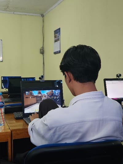 An individual is depicted from behind, seated at a wooden desk in what appears to be a computer lab or internet café in Muka, Indonesia. The person has short black hair, wears a light-colored long-sleeved collared shirt and dark trousers, and is seated on a black chair with a blue trim. They are actively engaged with a grey laptop displaying a first-person shooter video game, with their right hand positioned on an external black keyboard. A plastic bottle of "Aqua" brand water is on the desk to the left. A small black webcam is mounted atop the laptop screen. In the background, additional computer workstations are visible, featuring dark monitors and keyboards. One monitor to the right also has a webcam and displays text. The room features pale yellow walls with a calendar and an electrical cable running vertically. The ceiling is white.
