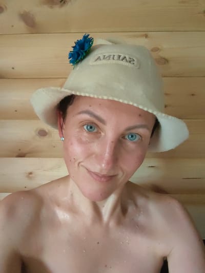 A woman with bright blue eyes and freckled skin is wearing a white sauna hat decorated with a blue flower. The hat has the word "SAUNA" embroidered on it in grey lettering. She is looking at the camera and smiling slightly, with a hint of sweat visible on her skin. The background is a wooden wall. The scene appears to be in a sauna in the rural area of Fanipalski Sel'ski Savet, Belarus.