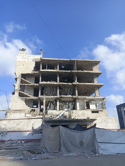 The media file shows a severely damaged multi-story building in an urban setting, likely in Gaza. The building's structure is heavily compromised, with missing floors, walls, and exposed concrete and rebar. Many of the concrete slabs that formed the floors are tilted or have collapsed, creating an uneven and hazardous appearance. The exterior walls are made of light-colored bricks and are riddled with numerous small holes, indicative of shelling or gunfire.
On the roof of the damaged building, three large loudspeakers are visible, pointing outwards. To the right of these, a smaller antenna structure is also present. The building's facade has sections with white and red horizontal stripes, remnants of its former state. A large, tattered grey tarp is stretched across the lower part of the building, possibly for temporary shelter or to cover damaged areas. Debris, including rubble and scattered pieces of construction material, litters the ground in front of the building and within its lower levels. To the right of the main damaged structure, a section of a more intact, darker-colored building can be seen, suggesting a contrast between areas affected by conflict and those that may be less so.
The sky above is a vibrant blue with scattered white clouds, suggesting it is daytime. The overall scene conveys a sense of destruction and the aftermath of conflict. No people are visible in the media file. There is some writing on the white brick wall of the damaged building, but it is difficult to decipher clearly.