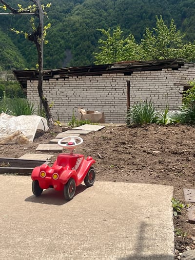 The image depicts an outdoor scene in Srednogortsi, Bulgaria. A red toy car is positioned on a concrete surface adjacent to a dirt area. A brick wall with visible structural damage is present, and a vine is growing nearby. In the background, a lush green hillside or mountain is visible. The scene appears to be taken during daylight hours. No people are visible in the image.