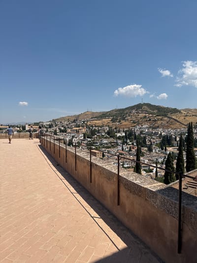 This image captures a panoramic view of the city of Granada, Spain, from an elevated vantage point, likely from the walls of the Alhambra. In the foreground, a terracotta brick walkway stretches along a weathered stone wall topped with metal railings. Several people are present on the walkway, some walking and others conversing. A man in a blue shirt and khaki shorts walks away from the camera on the left. Two individuals stand near the railing, seemingly engaged in conversation. Further along the walkway, another person stands near the railing.
Beyond the walkway and wall, the city of Granada unfolds. White-washed buildings with terracotta roofs are densely packed, cascading down hillsides. Numerous tall, slender cypress trees are interspersed throughout the urban landscape, adding vertical accents. In the background, a larger, dry, scrub-covered hill rises, topped with structures and antennas. The sky is a clear, bright blue with a few scattered white clouds. The sunlight casts long shadows on the brick walkway, indicating a sunny day. The overall scene is one of a historic European city bathed in warm, natural light.