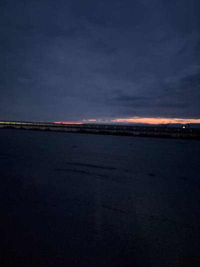 The image depicts a twilight landscape, likely at dusk, with the horizon line spanning horizontally across the middle. The upper half of the frame shows a vast, dark blue-gray sky, indicative of evening. Subtle cloud formations are visible within this expanse.
Along the distant horizon, a narrow band of warm light, featuring shades of orange and red, stretches across the scene. This illumination suggests either the final glow of a setting sun or a line of artificial lights from a distant feature. A single, distinct, bright yellow light source is visible within this illuminated strip on the horizon. The horizon itself is defined by a dark, linear element, possibly a distant bridge, roadway, or coastline, with subtle structural details that hint at guardrails or similar features.
The lower half of the image is dominated by a very dark, almost black, expanse. This area appears to be a body of water, such as a lake or sea, its surface rendered indistinct due to the low light conditions. The scene evokes a sense of calm and vastness at the transition from day to night. There is no visible text in the image. The location context is Cherna Gora, Bulgaria.