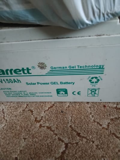 This image shows a close-up view of a white battery, likely a solar power gel battery, with green text and graphics. The text indicates it is a "Barrett V150Ah" and features "German Gel Technology". There are also caution warnings regarding short circuits, charging in sealed containers, sparks, and recharging after discharge. Recycling symbols for lead are visible, along with a "CE" mark. The battery is placed on a textured brown surface, possibly carpet or a similar material. The background is out of focus, showing a white and blue object, potentially fabric or plastic sheeting. There are no people visible in the image, and the environment appears to be indoors. The specific location in Hlushky, Ukraine is not discernible from the visual information presented.