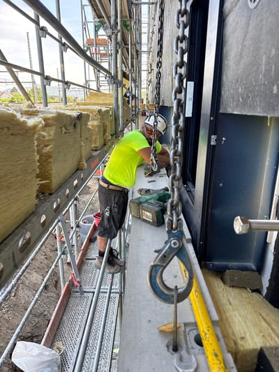 The image depicts a construction site in Rupperswil, Switzerland, likely part of a building renovation or new construction project. A worker, wearing a high-visibility yellow shirt, dark shorts, and a white hard hat with safety glasses, is actively engaged in work. He is positioned on a scaffolding platform, leaning over and appearing to be using a power drill.

The platform is constructed of metal scaffolding and planks, providing access to the upper levels of the building. Large, thick blocks of yellowish insulation material are stacked and secured along the exterior wall, suggesting insulation work is in progress. A chain hoist hangs prominently in the foreground, with a blue hook attached, indicating the potential for lifting heavy materials.

Tools and equipment are scattered on the platform, including a green power drill with batteries, a measuring tool, and a red bucket. The background shows more scaffolding, other construction materials, and a glimpse of the sky and surrounding buildings, characteristic of an urban or suburban construction environment. The overall scene conveys a busy and active construction setting.