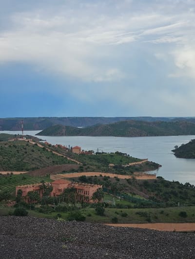The image presents an elevated panoramic view of a landscape in Oumazza, Morocco, featuring a large body of water surrounded by undulating terrain. The foreground consists of a dark, coarse gravel or rocky surface. In the midground, a substantial, multi-story building complex with terracotta-colored facades and roofs, displaying fortress-like architectural elements including crenellations, is situated amidst green vegetation and numerous palm trees. Adjacent to this complex, a winding dirt path or road is discernible, along with a long, light brown retaining wall that follows the contours of the hillside towards the water. The landscape includes terraced hillsides and scattered patches of green and brown vegetation.
The central feature of the scene is a vast, calm body of water, likely a reservoir or lake, which extends through the middle and background, curving between several green and arid hills. On a distant hill to the left, a tall, slender communications tower with alternating red and white bands is visible. Several smaller buildings with terracotta roofs are sparsely distributed on the hillsides nearer to the water. The sky is partially overcast, exhibiting a gradient from light grey to muted blue, with diffuse cloud cover. No individuals or specific actions are discernible within the frame.