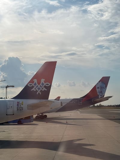 Two Serbian Air Force Airbus A330-200 aircraft are parked on the tarmac at Belgrade Nikola Tesla Airport in Surcin Urban Municipality, Serbia. The tail fin of the aircraft on the left displays a stylized blue and white emblem against a red background, with the text "EXPO 2027" and "BEOGRAD SRBIJA" printed on the fuselage. The tail fin of the aircraft on the right features a portrait of Nikola Tesla against a red background. The aircraft are positioned at an angle, with the foreground dominated by the airport tarmac, marked with white lines and orange traffic cones. In the background, a large cumulonimbus cloud is visible in the sky, along with construction cranes on the left. The lighting suggests it is daytime, with shadows cast on the tarmac.