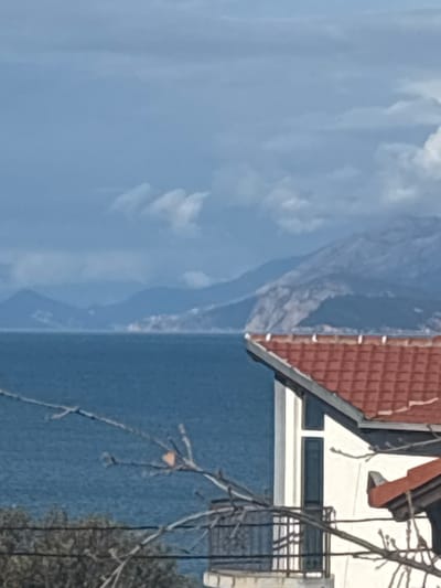 The scene is an exterior landscape shot from Utjeha-Bušat, Montenegro. The foreground is occupied by bare tree branches with a few dried, light-colored leaves and dark utility wires running horizontally across the lower-middle section of the frame. To the right, a portion of a white building with a red-orange terracotta tiled roof is visible. This building features a vertical window section and a balcony enclosed by a black metal railing. A second, lower roof structure is partially discernible beneath the main building on the right. The midground consists of a broad expanse of dark blue water, consistent with the Adriatic Sea, exhibiting slight surface agitation. The background is composed of a distant chain of rugged, blue-grey mountains forming a coastline, with a prominent light-colored cliff face observed on the right segment of the mountain range. The sky above is overcast with a mix of light and dark grey clouds. No individuals or discernible activities are present.