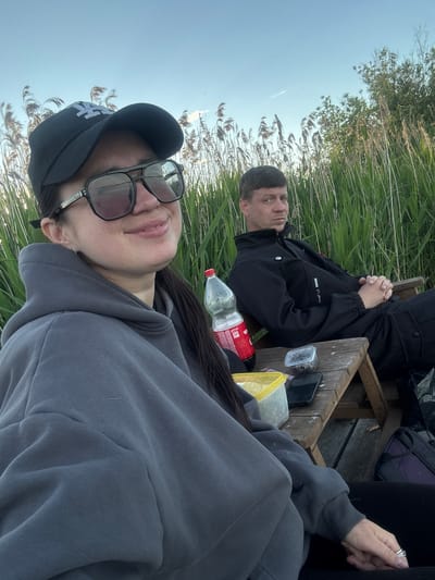 Two individuals are outdoors, reclining near tall grasses and reeds. One person, wearing a gray hooded sweatshirt and a baseball cap with sunglasses perched on the cap's brim, is in the foreground. Behind them and partially obscured by a wooden surface with assorted items (including a bottle, containers, and a mobile phone), the other person is dressed in a black jacket. The setting appears to be a natural environment, possibly near a body of water. The time of day is likely late afternoon or early evening, as indicated by the ambient light. The precise location within Khryashchevka, Russia cannot be determined from the image alone. The scene appears to depict a moment of leisure.