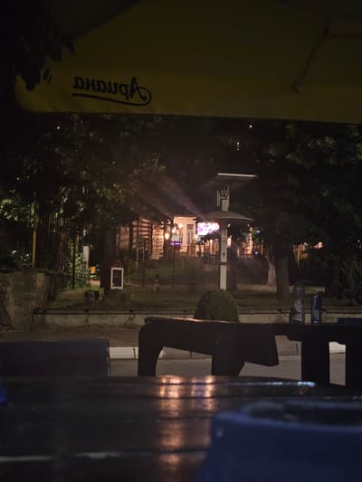 The image depicts an outdoor seating area at night, possibly a cafe or restaurant, with a yellow awning overhead featuring dark lettering. In the background, a building with illuminated windows is visible, surrounded by trees and a wooden fence. A small, red and white structure resembling a mailbox or information booth is positioned in front of a tree. In the foreground, dark-colored benches and tables are present, with reflections of light on their surfaces. Two bottles, one clear and one blue, are placed on a table in the middle ground. The scene suggests a dimly lit, tranquil evening setting. The location appears to be in the city of Byaga, Bulgaria.
