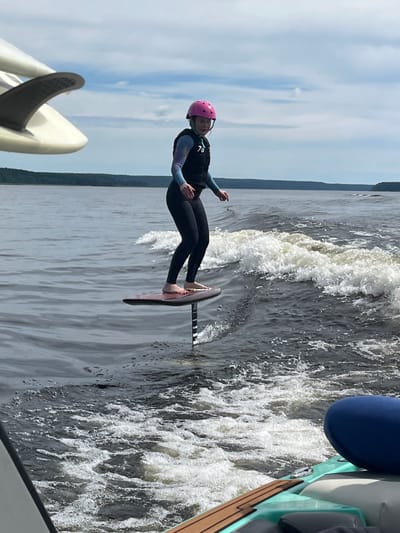 A young person wearing a pink helmet and a black wetsuit with colorful sleeves is hydrofoiling on a body of water. They are standing on a hydrofoil board, which is a board with fins that lifts out of the water as it gains speed. The person's feet are visible, and they appear to be in the process of riding. The water is dark and choppy, with waves breaking around the board. In the foreground, part of a boat is visible, with a blue cushion and wooden deck. The background shows a distant shoreline with trees under a cloudy sky. The scene is outdoors and appears to be a sunny or partly cloudy day. The location is identified as Gromovo, Russia.