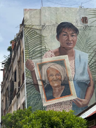 This is a close-up, low-angle shot of a mural on the side of a weathered, multi-story building. The mural depicts a woman holding a framed photograph of an elderly woman. The main figure in the mural, a woman with dark hair pulled back and a light blue scarf draped over her shoulder, looks directly at the viewer. She is wearing a patterned dress. The framed photograph, held in her hands, shows an elderly woman with a white headscarf, her face lined with age and a gentle smile. Both the main mural and the photograph are rendered in a realistic style.
The building itself shows signs of age, with peeling paint, exposed brick in one section, and some visible metal fixtures and wires attached to the wall, possibly for bird deterrence. Greenery, likely trees or vines, is visible growing from ledges of the building and in the foreground, obscuring the lower portion of the building and the street below. The sky above is overcast and pale blue. Red spray paint graffiti, including a date that appears to be "2025", is visible on the upper right portion of the building's facade.
This mural is located on a building on Street 2, in the heart of Battambang, Cambodia, a city renowned for its vibrant street art scene. The artwork is part of a larger collection of murals in the city, often depicting local life and history.