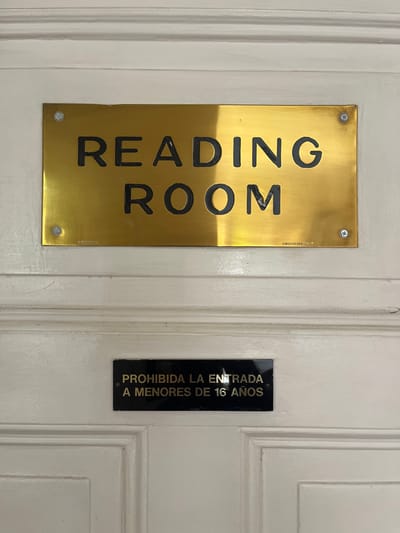 A white door, likely made of wood, is depicted in a close-up, centered vertical shot within a building in Hurlingham, Argentina. Two rectangular signs are affixed to the door. The upper sign is a gold-colored metal plaque, secured by four visible screws, displaying the phrase "READING ROOM" in prominent black capital letters. Small, illegible text appears in the bottom left and right corners of this plaque. A horizontal shadow line bisects the gold sign. Below this, approximately centered on a lower panel of the door, is a smaller, black rectangular sign with the gold capital text "PROHIBIDA LA ENTRADA A MENORES DE 16 AÑOS," which translates to "ENTRY FORBIDDEN TO PERSONS UNDER 16 YEARS OLD." The door features subtle decorative molding, indicating paneled construction, and exhibits minor surface wear and marks. No individuals are visible. The environment consists solely of the door and its attached signage.