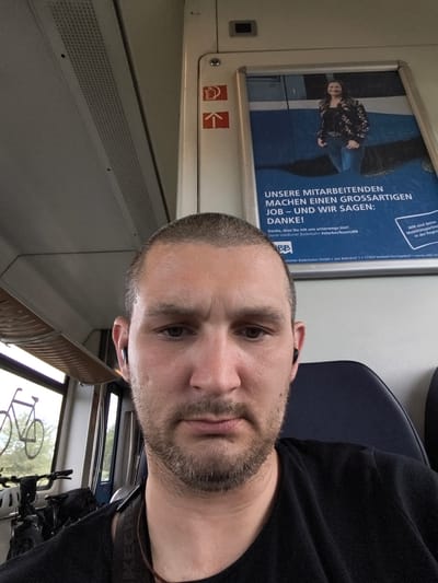 The image is a selfie taken inside a train. The man in the foreground has short, dark hair, a light beard, and is wearing a black t-shirt and black earbuds. In the background, a poster is visible, featuring a smiling woman in a floral jacket and jeans, with German text that translates to "Our employees make a great job - and we say: Thank you!". The poster also includes the logo "BB" and text related to Usedomer Bäderbahn. The train interior shows rows of seats and windows. Through the window, bicycles are visible, suggesting a bike-friendly transport option. The location is identified as Züssow, Germany.