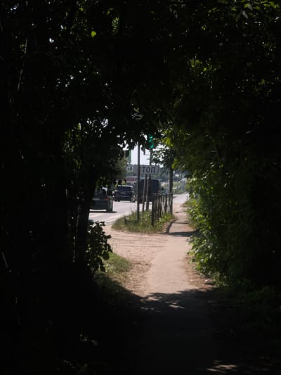 The image depicts a shaded pathway leading towards a street, bordered by dense foliage on both sides. The pathway itself appears to be made of gravel or dirt, with some patches of grass growing along its edges. Sunlight filters through the leaves, creating dappled patterns on the ground and casting shadows.
In the distance, a street is visible, with a few vehicles present. One vehicle, a black SUV, is stopped at what appears to be a stop sign. Next to the stop sign, a traffic light is visible, showing a green light. Another vehicle, a lighter colored SUV, is also present on the street.
The scene suggests a rural or suburban setting in Russia, given the context of "Юдино, Russia." The time of day appears to be daytime, likely late morning or early afternoon, given the bright sunlight. The weather seems to be clear and sunny.
The overall mood is somewhat tranquil, with the natural elements of the trees and foliage creating a sense of enclosure and shade. The presence of the vehicles and traffic signals indicates a transition from a more natural environment to a developed area.
The visible text on the sign reads "СТОП," which is the Russian word for "STOP."