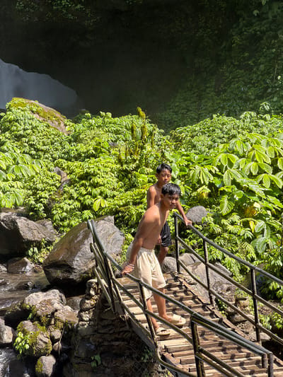 Two shirtless boys, one in black shorts and the other in light-colored shorts and sandals, stand on a wooden staircase at a waterfall in Sawan, Indonesia. The staircase, made of wooden planks and metal railings, descends towards a rocky stream. Lush green vegetation covers the surrounding rocks and cliffs, with a large rock overhang obscuring the top of the waterfall. The water from the waterfall creates a misty effect in the dark cave-like area behind the boys.