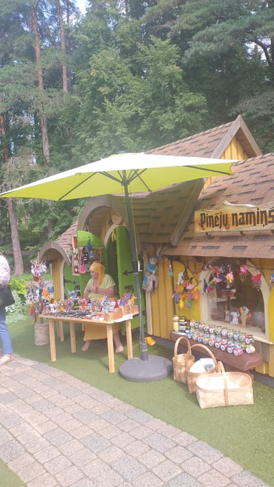 The image depicts an outdoor marketplace stall, potentially located in Kroņauce, Latvia, as suggested by the sign "Pineļu namins". A woman, wearing a yellow headscarf and a light green top, is seated behind a wooden table laden with an assortment of handmade crafts, including small stuffed animals and decorative items. The stall itself is a whimsical, miniature-house-like structure painted yellow with brown shingled roofs. A large, bright green umbrella is set up to provide shade over the stall and the woman. In front of the stall, a cobblestone path leads away from the viewer, and to the right, several woven baskets are displayed on the artificial turf ground. The background consists of lush green trees, suggesting a natural or park-like setting. The scene appears to be a sunny day, with a relaxed atmosphere at the marketplace.