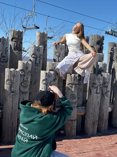 A photo taken in Gubakha, Russia shows a young female, with light brown hair, wearing a white t-shirt and long white and beige skirt, posing on a structure made of carved wooden logs, while another person in a green hoodie and black baseball cap kneels to photograph her. The backdrop features a clear blue sky and a ski lift structure in the distance. The carved logs have repetitive facial designs, adding a cultural or artistic dimension to the scene. A wooden platform is partially visible at the right edge.