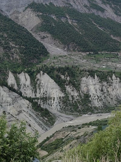 This image captures a broad view of a mountain valley in Gondhla, India. A wide, muddy river flows through the valley floor. Steep, rocky slopes rise on either side of the river, covered in patches of dense green trees. In the upper part of the image, terraced fields are visible on the mountainside, indicating agricultural activity. The overall scene is rugged and natural, with evidence of human settlement in the form of the terraced fields.