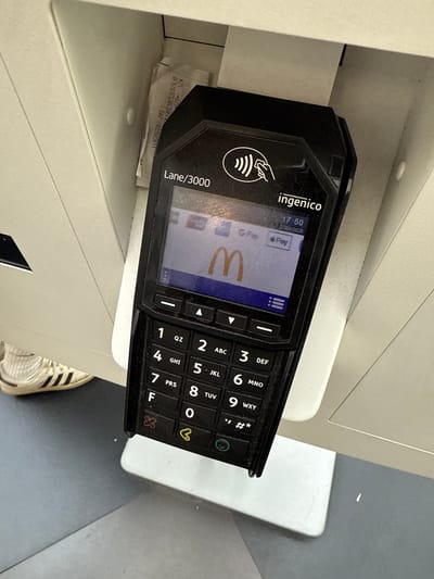 The image shows a close-up of a black Ingenico Lane/3000 payment terminal displaying the McDonald's logo. The terminal is positioned vertically and appears to be mounted on a light-colored structure, possibly part of a service counter. The screen shows "G Pay" and "Play" icons, indicating contactless payment readiness. A receipt is partially visible to the left of the terminal, showing some alphanumeric characters. Below the screen is a keypad with numbers 1 through 9, 0, and other symbols. Some colorful stickers are attached to the keypad: a red X on the 'F' key, a yellow heart on the '0' key, and a green circular button at the bottom. A portion of a white and black striped sneaker is visible in the lower left corner of the image. The background is a neutral, indoor setting with muted lighting. The scene is indoors, in what appears to be a public establishment, possibly a McDonald's restaurant in Białystok, Poland.