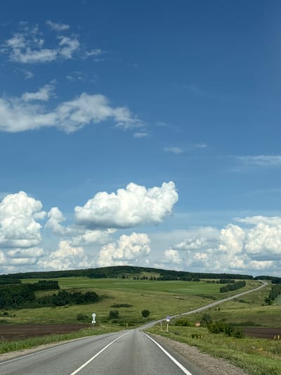 The image depicts a scenic view of a rural landscape in Chubovka, Russia, on a sunny day with scattered white clouds against a bright blue sky. A paved road stretches into the distance, winding through rolling green hills covered in fields and scattered trees. The foreground shows the asphalt road with white lane markings. On the left side of the road, there is a patch of dark brown soil, likely a field. Further back, the landscape is a vibrant green, with some areas appearing as cultivated fields and others as grassy meadows. A line of trees forms a dark green band along the crest of the hills in the middle ground. In the far distance, the hills are covered in a dense forest. There are a few small vehicles visible on the road in the distance. Two road signs are present on the right side of the road in the mid-ground. The overall impression is one of natural beauty and open space, characteristic of the Russian countryside.
