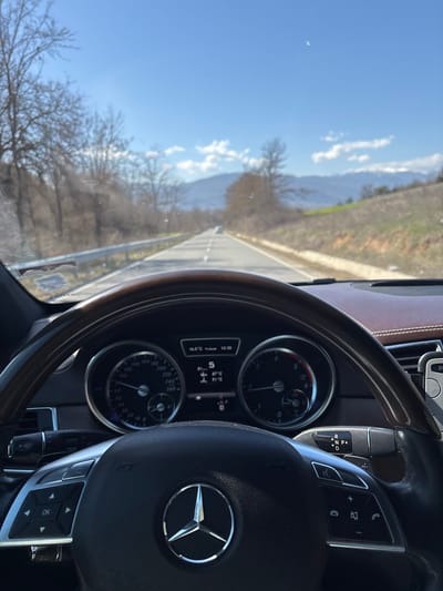 The image displays the interior of a Mercedes-Benz vehicle, viewed from the driver's perspective. The lower half of a black steering wheel with a central Mercedes-Benz logo is prominent, featuring silver control buttons on its left spoke. The upper rim of the steering wheel appears to be dark wood-grained. Beyond the steering wheel, the instrument cluster is visible, showing an ambient temperature of 16.0°C and the time as 13:30. The central digital display indicates the vehicle is in gear '5', traveling at 81 km/h, with additional temperature readings of 87°C and 91°C. A gear selector stalk is partially visible on the right, indicating 'P', 'R', 'N', 'D' positions. The dashboard is finished in a brown, possibly leather, material.
Through the windshield, a two-lane asphalt road stretches into the distance, marked with a broken white line for lane separation and a solid white line on the right shoulder. A silver metal guardrail lines the left side of the road. The environment surrounding the road consists of bare deciduous trees and dry, grassy embankments, indicating late winter or early spring conditions. In the far distance, hills or mountains with some lighter patches, possibly snow, are visible. The sky is clear blue with scattered white cumulus clouds, suggesting a sunny day. A single white car is observed in the distance on the road ahead. The scene is located in the city of Blatska, Bulgaria.
